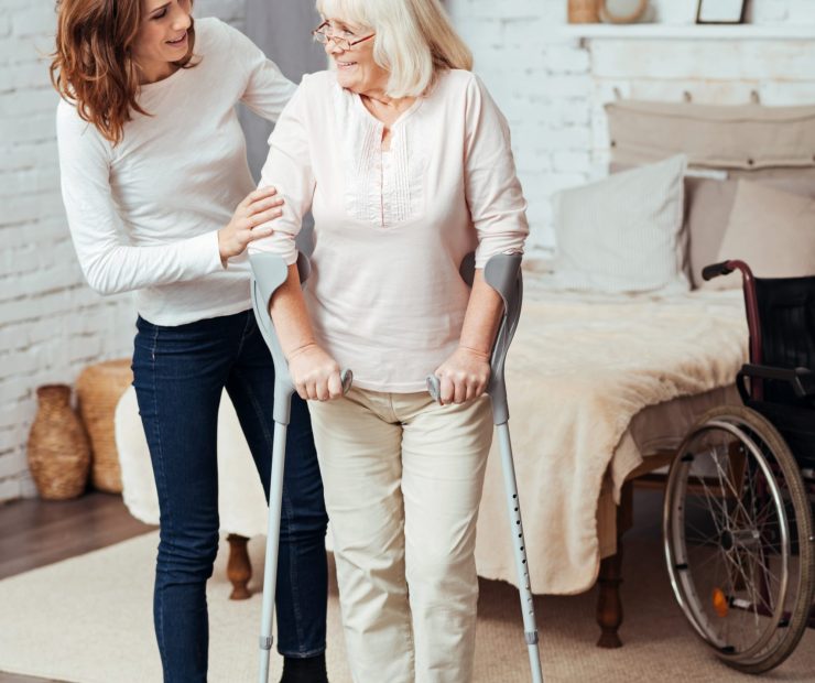 One more step. Pleasant caring young woman helping her grandmother to walk with crutches while providing rehabilitation at home